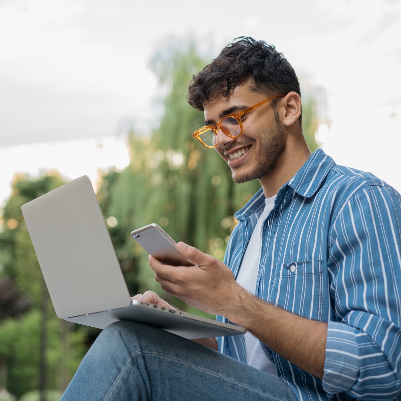 Man in a park holding his phone and his laptop, smiling