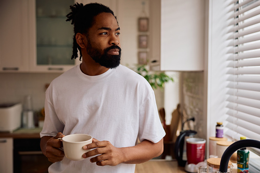 Man holding a coffee cup in his kitchen looking out the window. He looks contemplative.